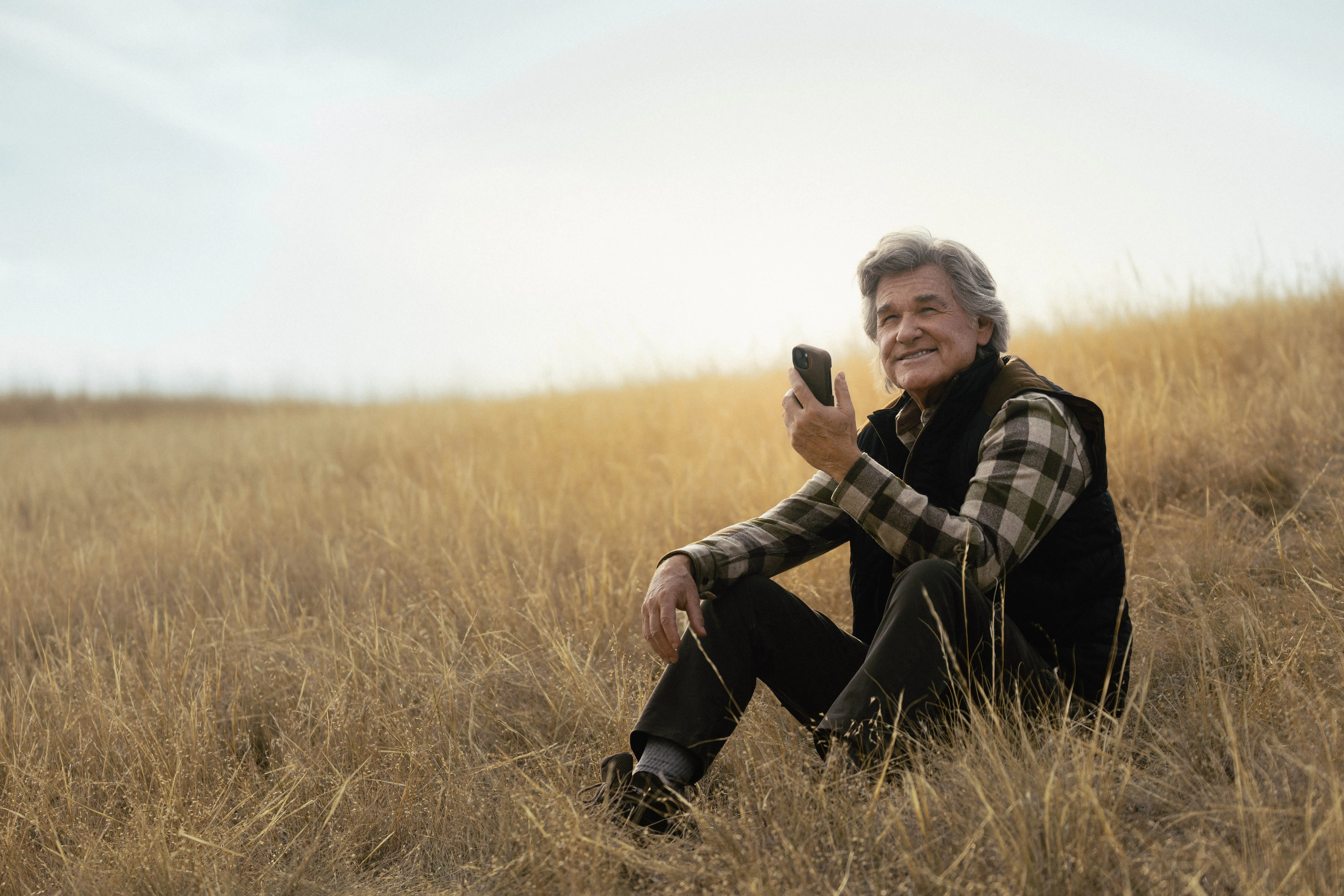 Preston Clyburn (Kurt Russell) smiling while holding a phone and sitting in a field of dry grass.