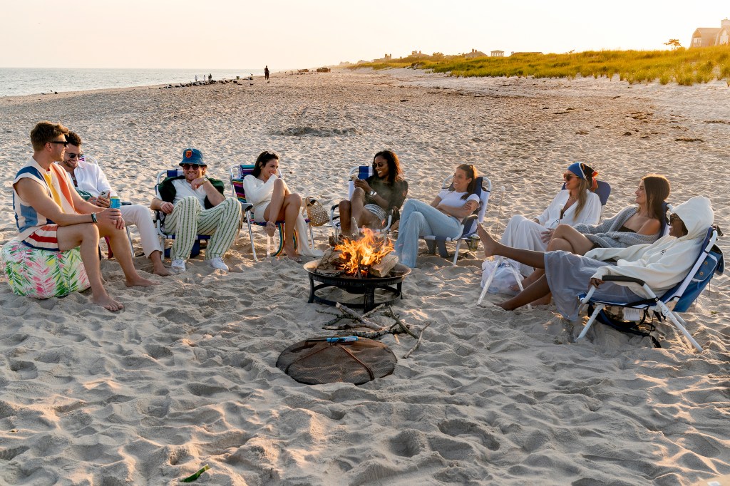 Kyle Cooke, Jesse Soloman, West Wilson, Paige DeSorbo, Ciara Miller, Amanda Batula, Lindsay Hubbard, Lexi Wood, and Gabby Prescod sitting around a bonfire on the beach at sunset.