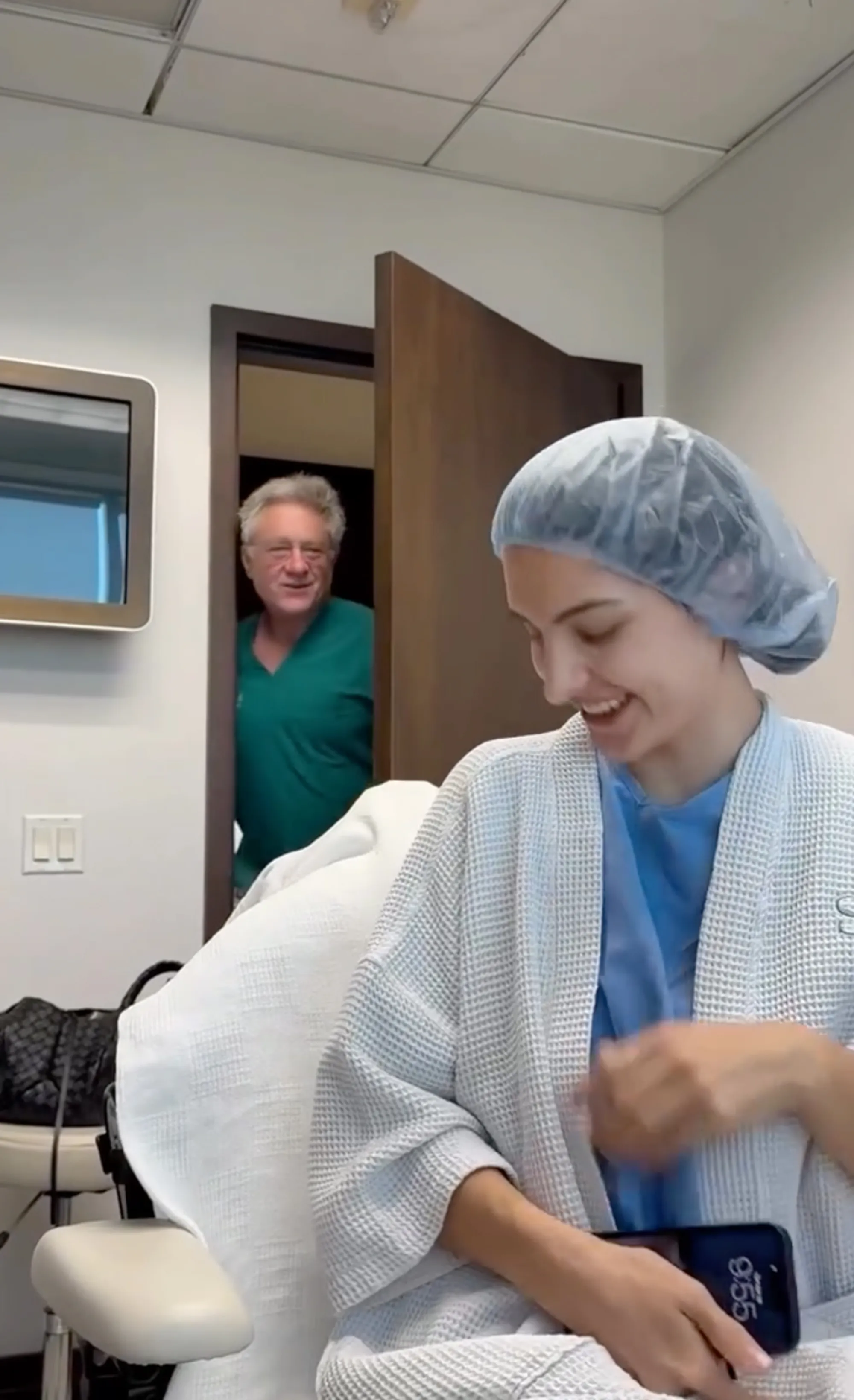 Sophia, Kyle Richards' daughter, in a hospital gown and hairnet, laughs in an exam room while a doctor in teal scrubs peeks through an open doorway.