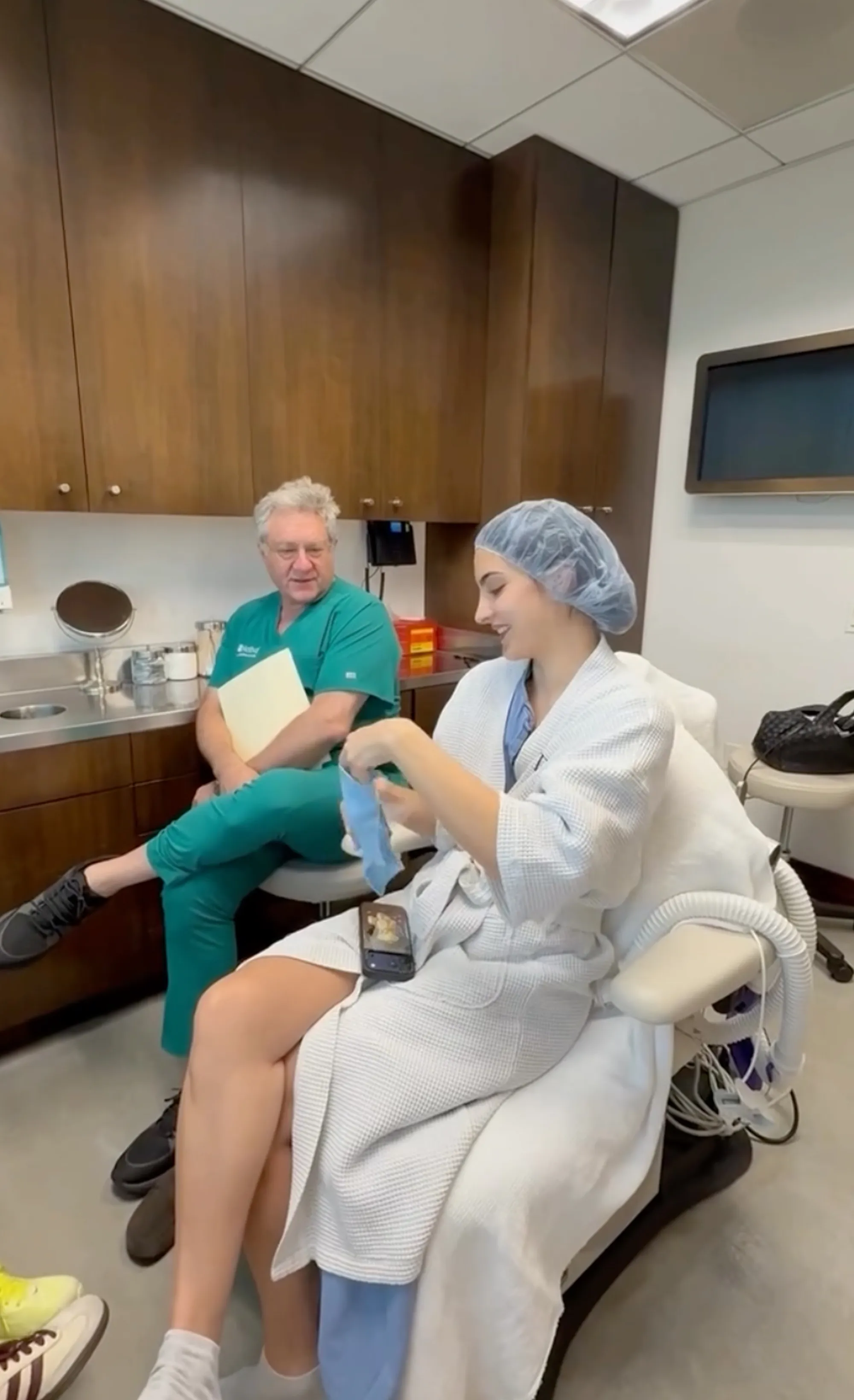 Sophia, Kyle Richards' daughter, in a hospital gown and hairnet, laughs in an exam room while a doctor in teal scrubs peeks through an open doorway.