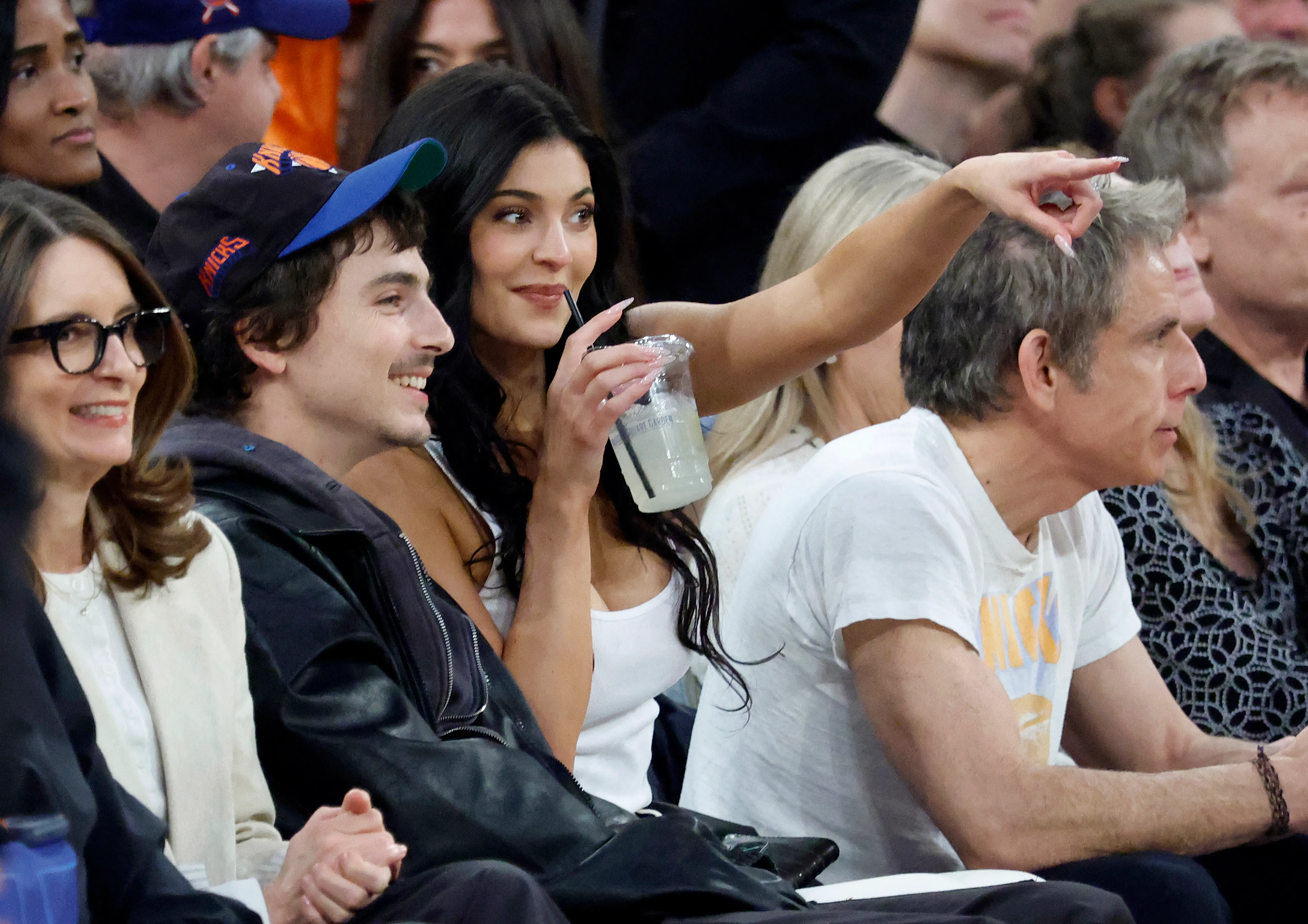 Kylie Jenner smiles while holding a drink and Timothée Chalamet wears a Knicks cap at a basketball game.