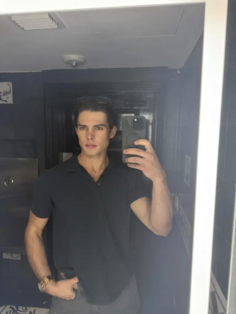 Man in a black polo shirt, black sunglasses, and a watch, taking a mirror selfie with a phone.