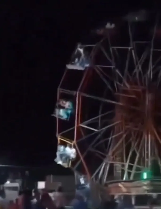 A large Ferris wheel with several occupied gondolas appears to be tilting or collapsing at a night fair.