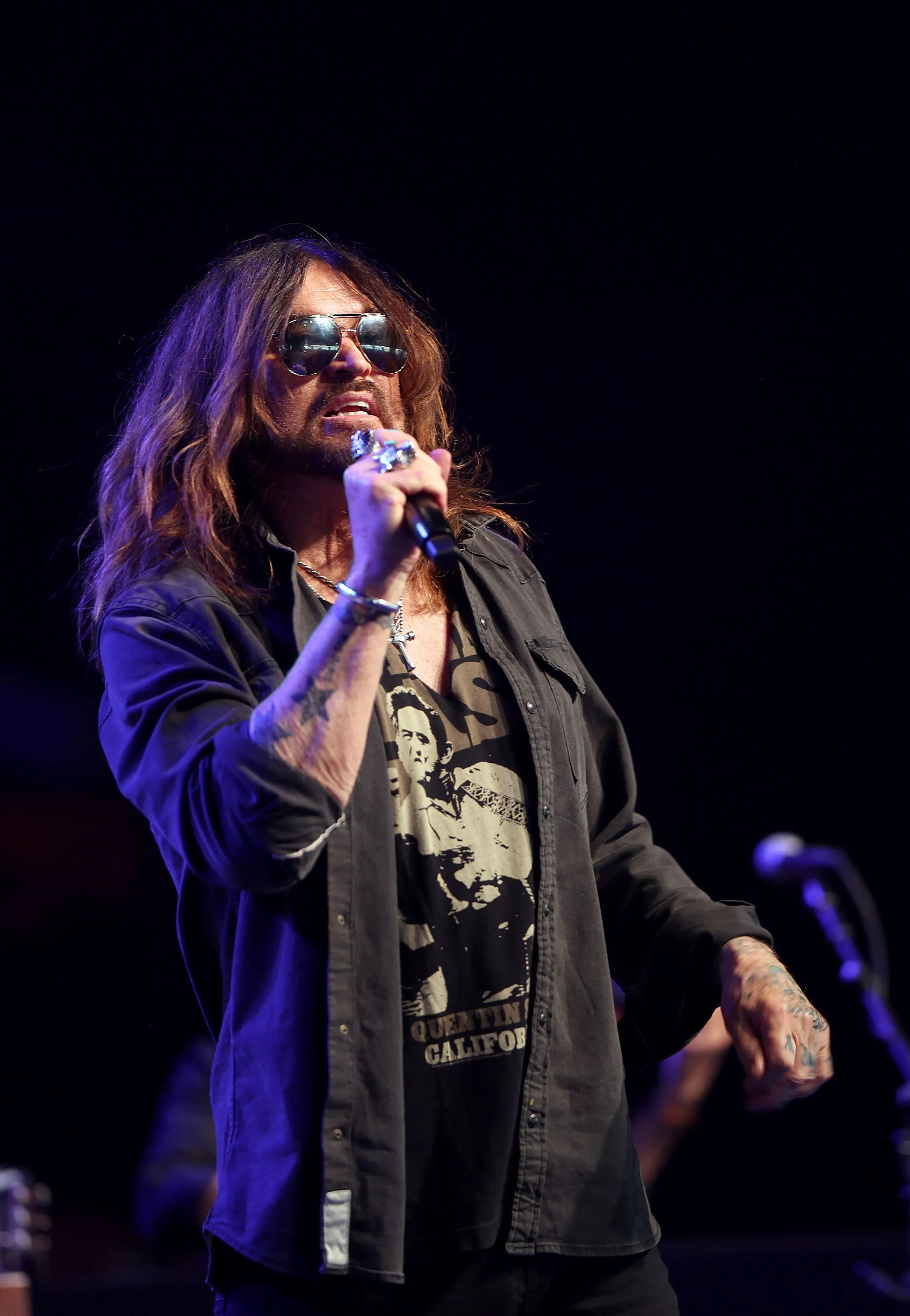 Billy Ray Cyrus singing into a microphone on stage, wearing sunglasses, a dark shirt, and an open dark overshirt.