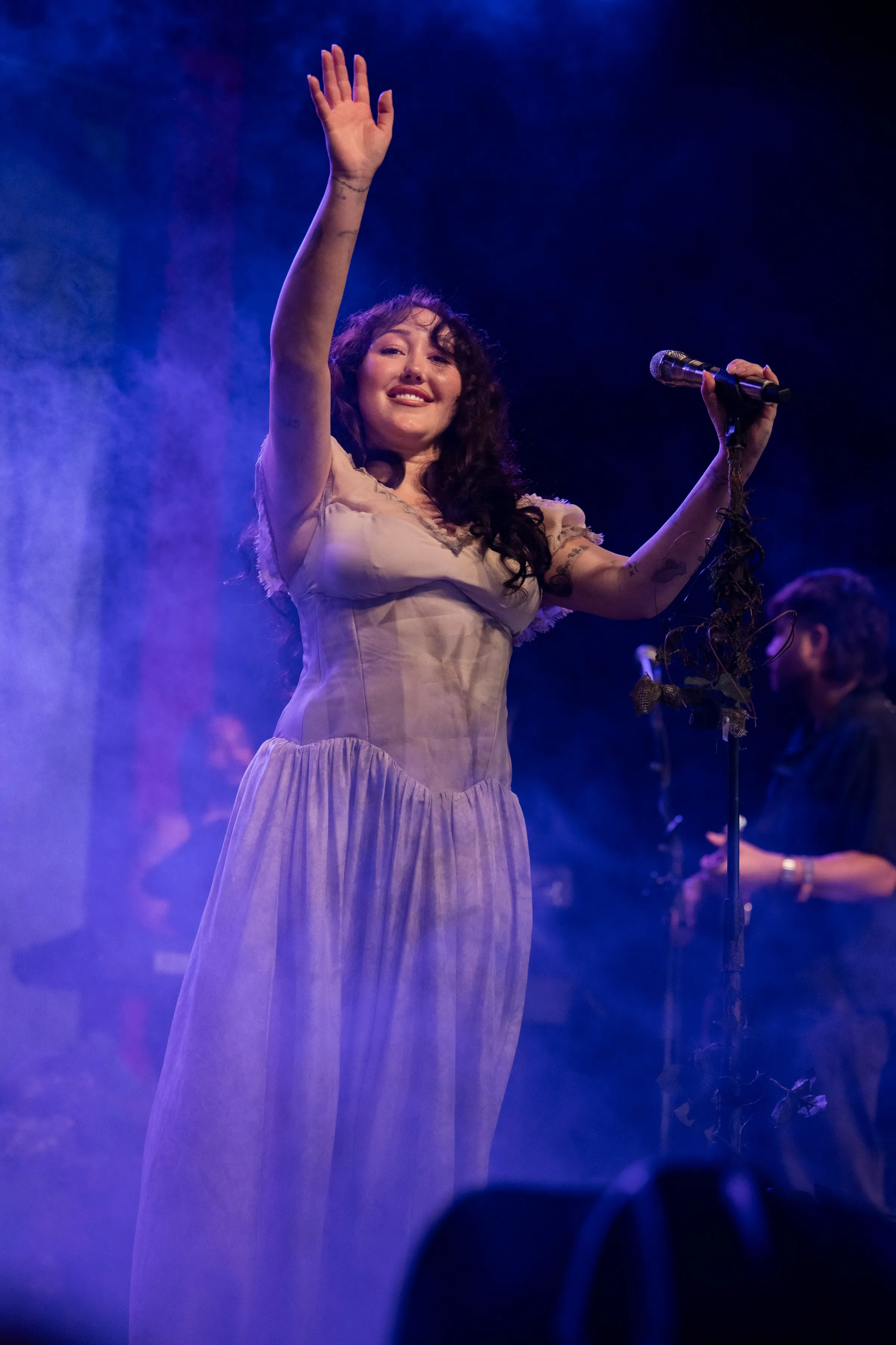 Noah Cyrus on stage, raising her right hand, holding a microphone in her left hand, wearing a flowing dress under blue and purple stage lights.