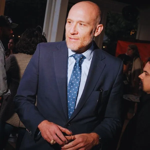 A man in a suit with a blue tie, a bald head, and a short beard, wearing a ring, looks to his right at an event.