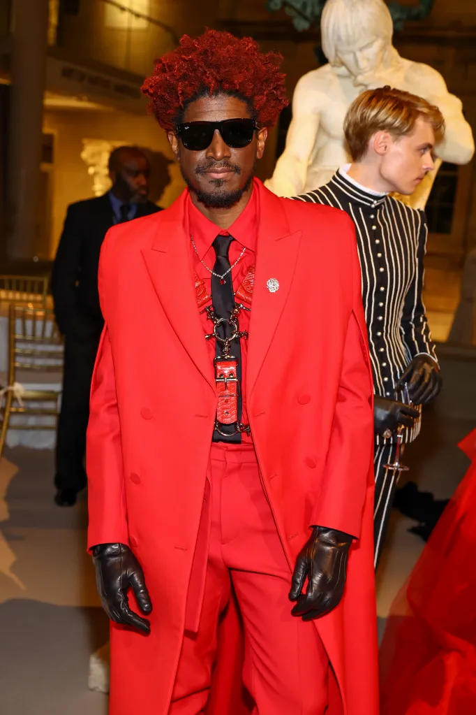 Labrinth wearing a red suit, black tie, black gloves, and sunglasses, with a man in a striped jacket and a statue behind him at the Met Gala.