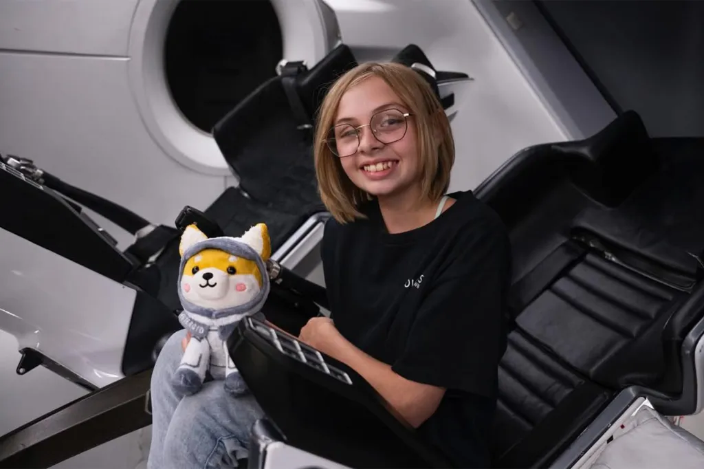 Liv Perrotto, a young girl with blonde hair and glasses, smiles while holding a plush toy in what appears to be a SpaceX spacecraft.