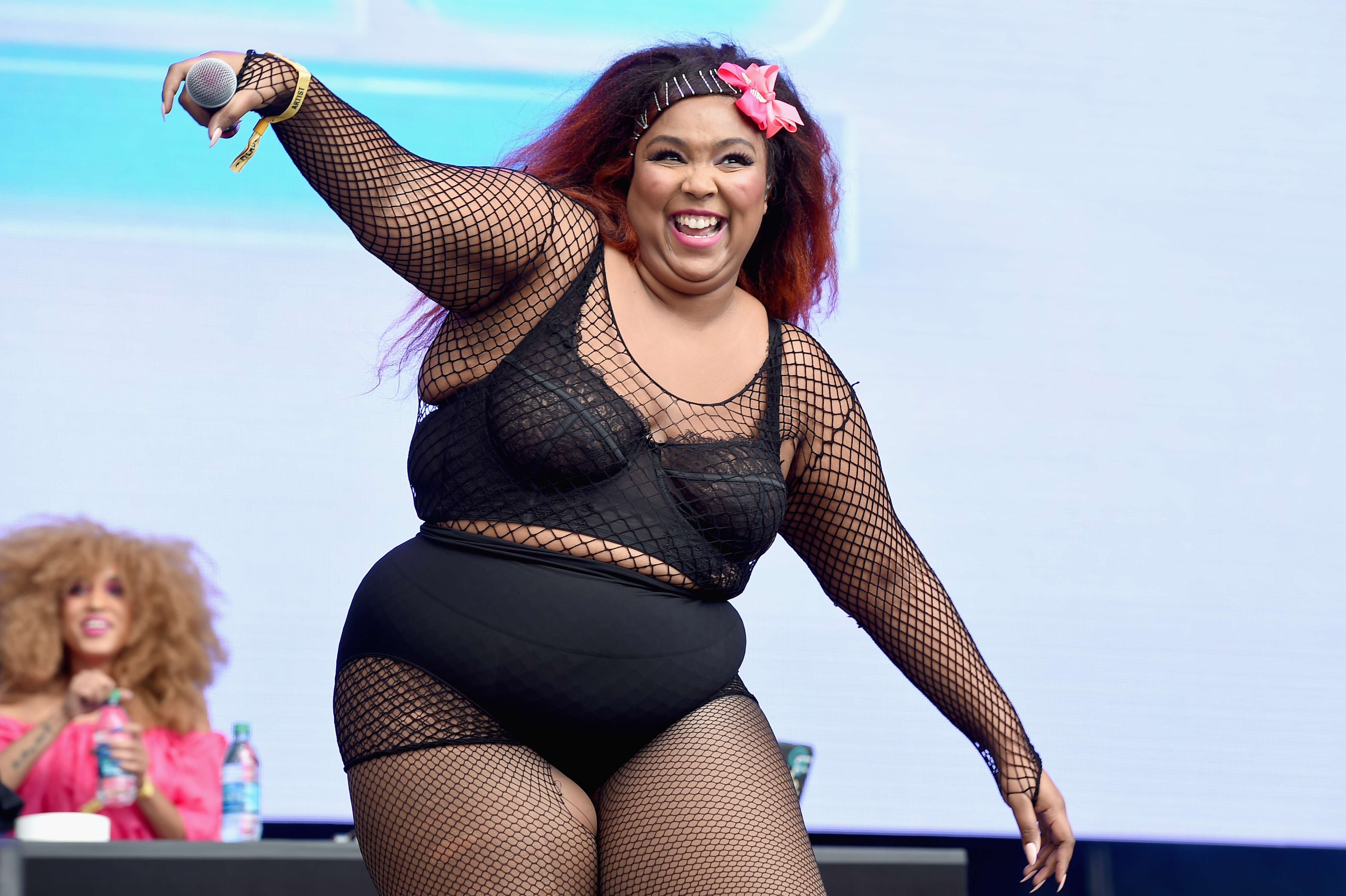 Lizzo performing onstage wearing a black lace bra and fishnet bodysuit, holding a microphone.