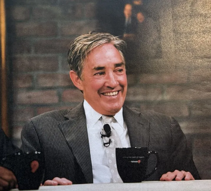 Raymond P. Hartmann smiling while wearing a suit and tie, with a black mug in front of him.