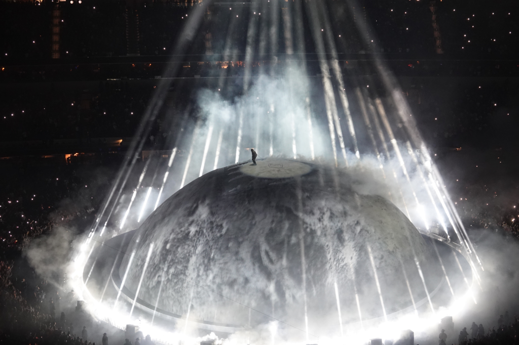 Kanye West performs on top of a moon-like stage, surrounded by spotlights and smoke, during a concert at SoFi Stadium.