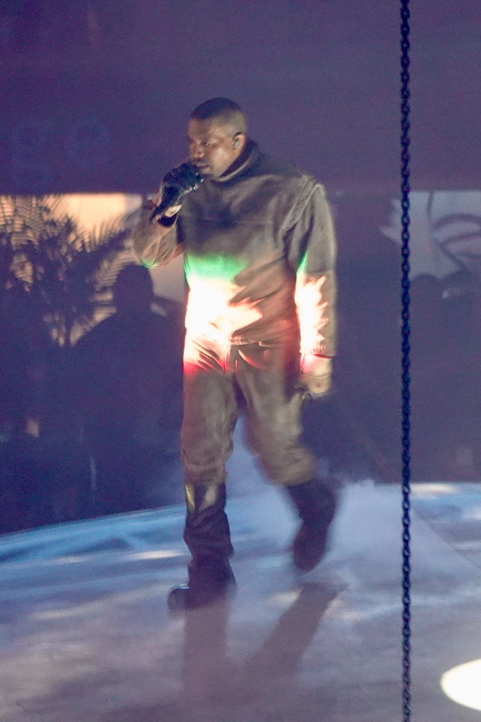 Kanye West performing on stage, wearing a gray outfit and holding a microphone.
