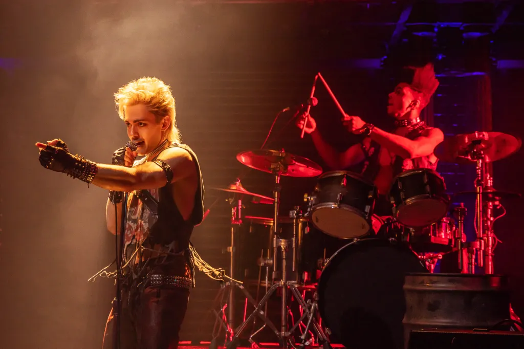 Ali Louis Bourzgui singing while pointing, with Dean Maupin playing drums in the background on the set of 