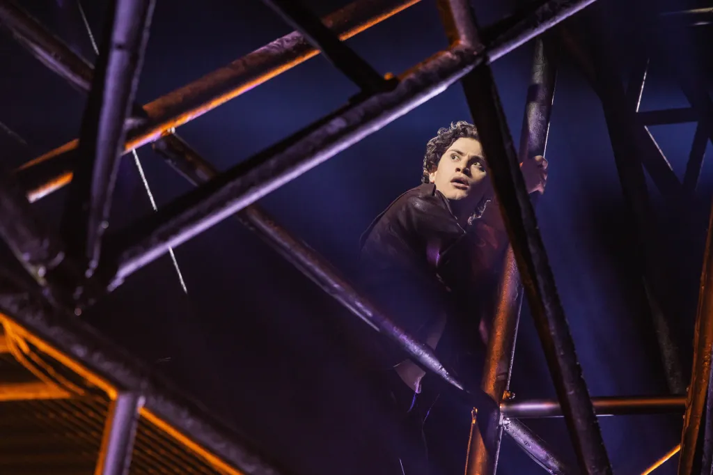 LJ Benet as a Lost Boy on Broadway, peering through metal scaffolding.