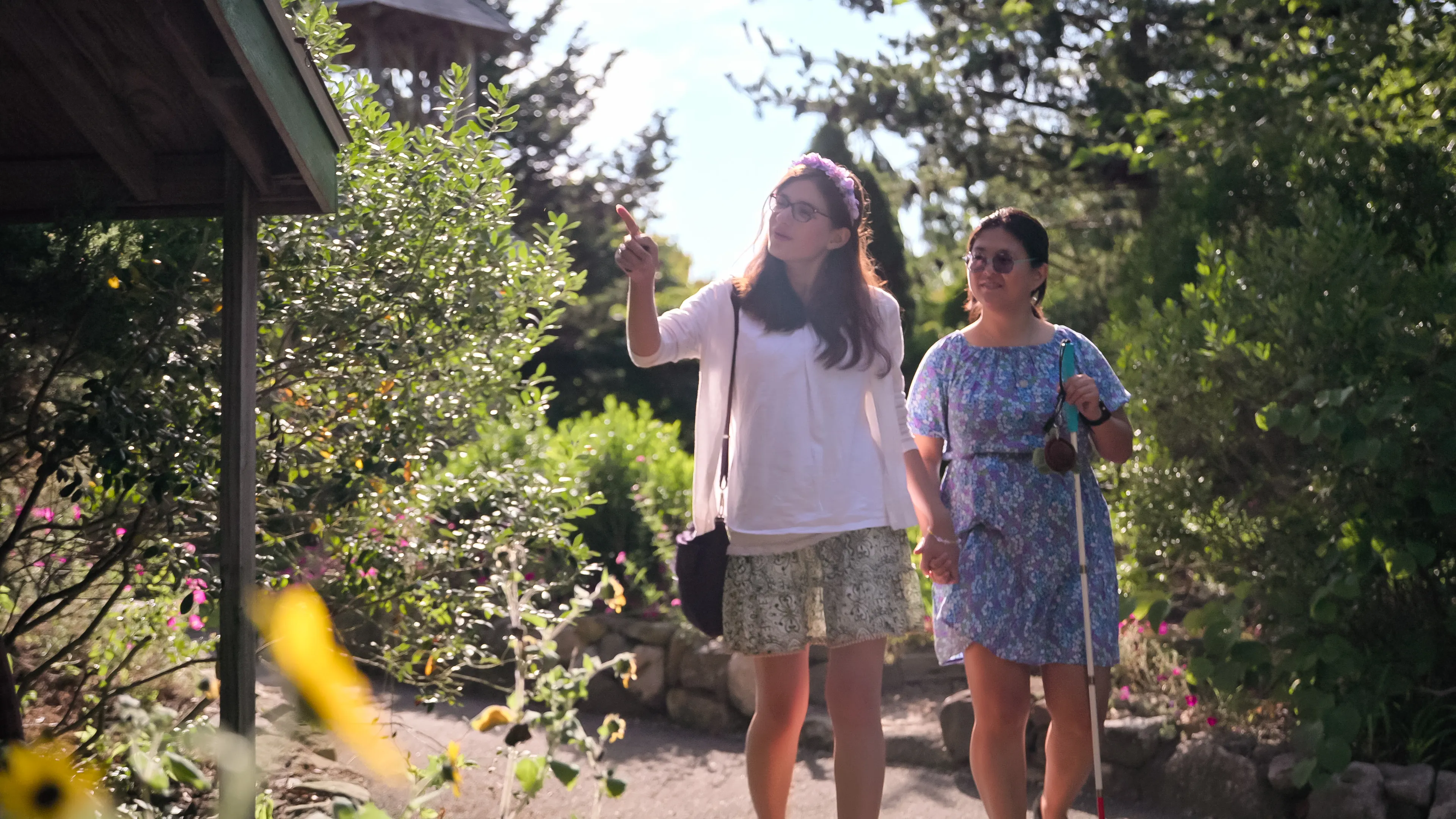Two women walk hand-in-hand through a garden, with one woman pointing to something off to the left.