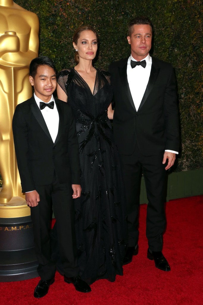 Angelina Jolie in a black gown, Maddox Jolie-Pitt, and Brad Pitt in a black tuxedo pose on a red carpet.
