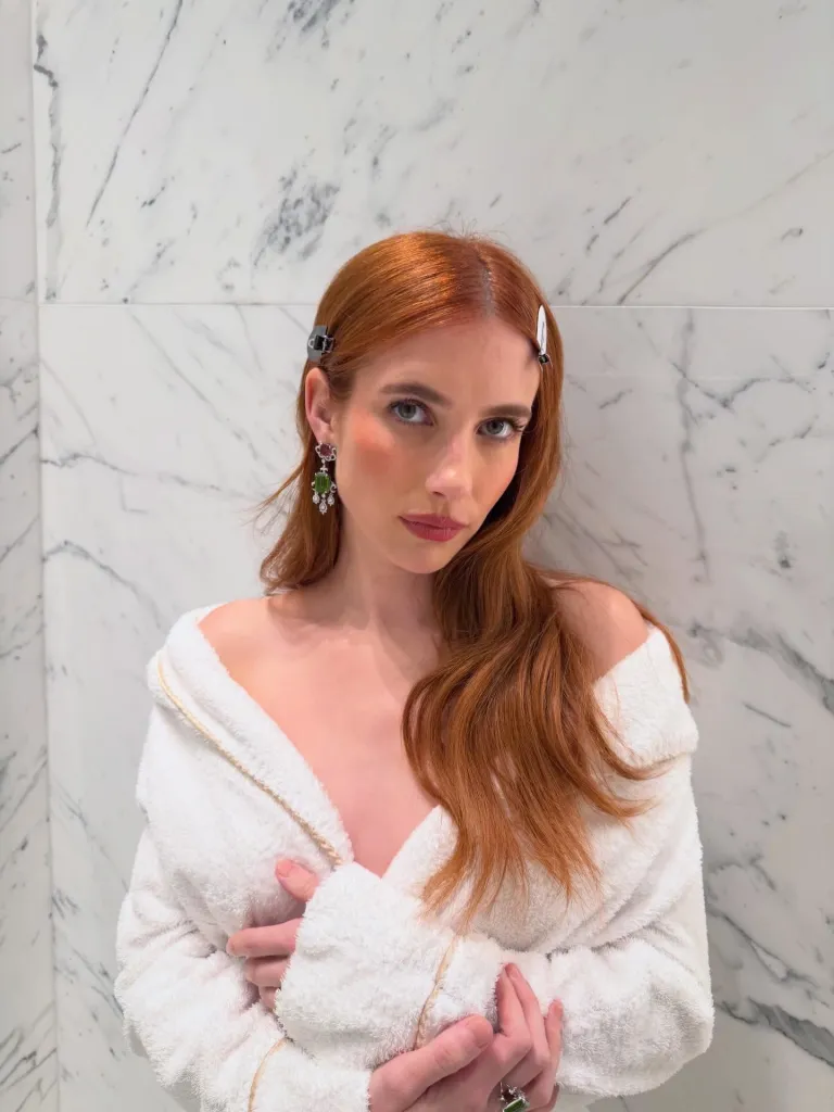Emma Roberts in a white robe with hair clips and emerald earrings.