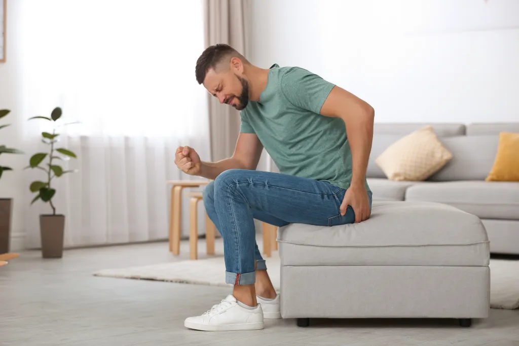 A man in jeans and a green shirt suffering from hemorrhoid pain while sitting on an ottoman in a living room.
