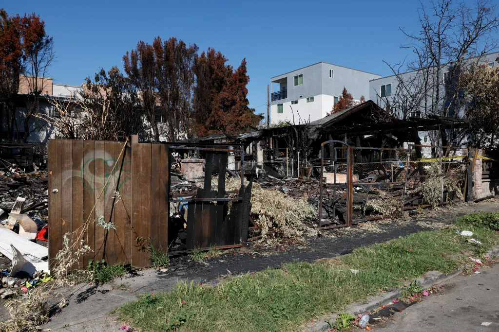 The remains of a burned house with charcoaled debris and a collapsed roof, next to a modern gray and white apartment building.