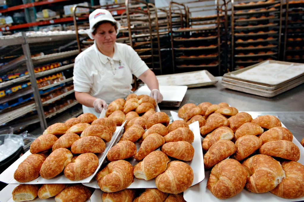 A Costco employee wearing a white shirt, hairnet, and hat, places trays of croissants onto a large metal table.