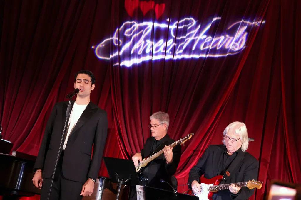 Matteo Bocelli sings into a microphone with two guitarists on stage, in front of a red curtain with the words 