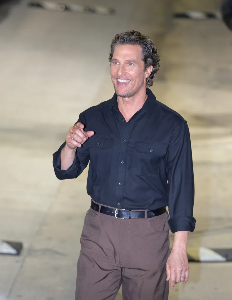 Matthew McConaughey wearing a black shirt and brown pants, pointing and smiling.