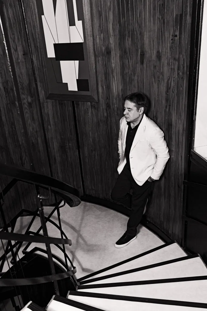 Maximilien de Hoop Cartier, also known as Max Cartier, standing on a winding staircase, leaning against a dark wood-paneled wall, looking down.