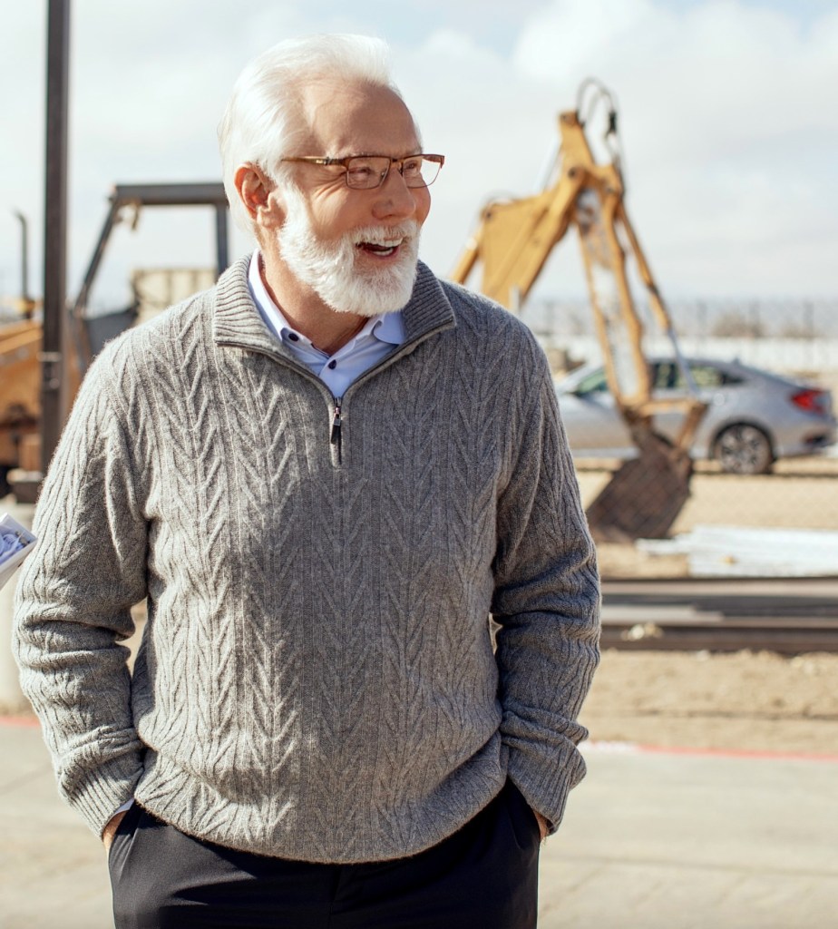 Mayor R. Rex Parris smiling, wearing a gray sweater and glasses, with construction equipment in the background.
