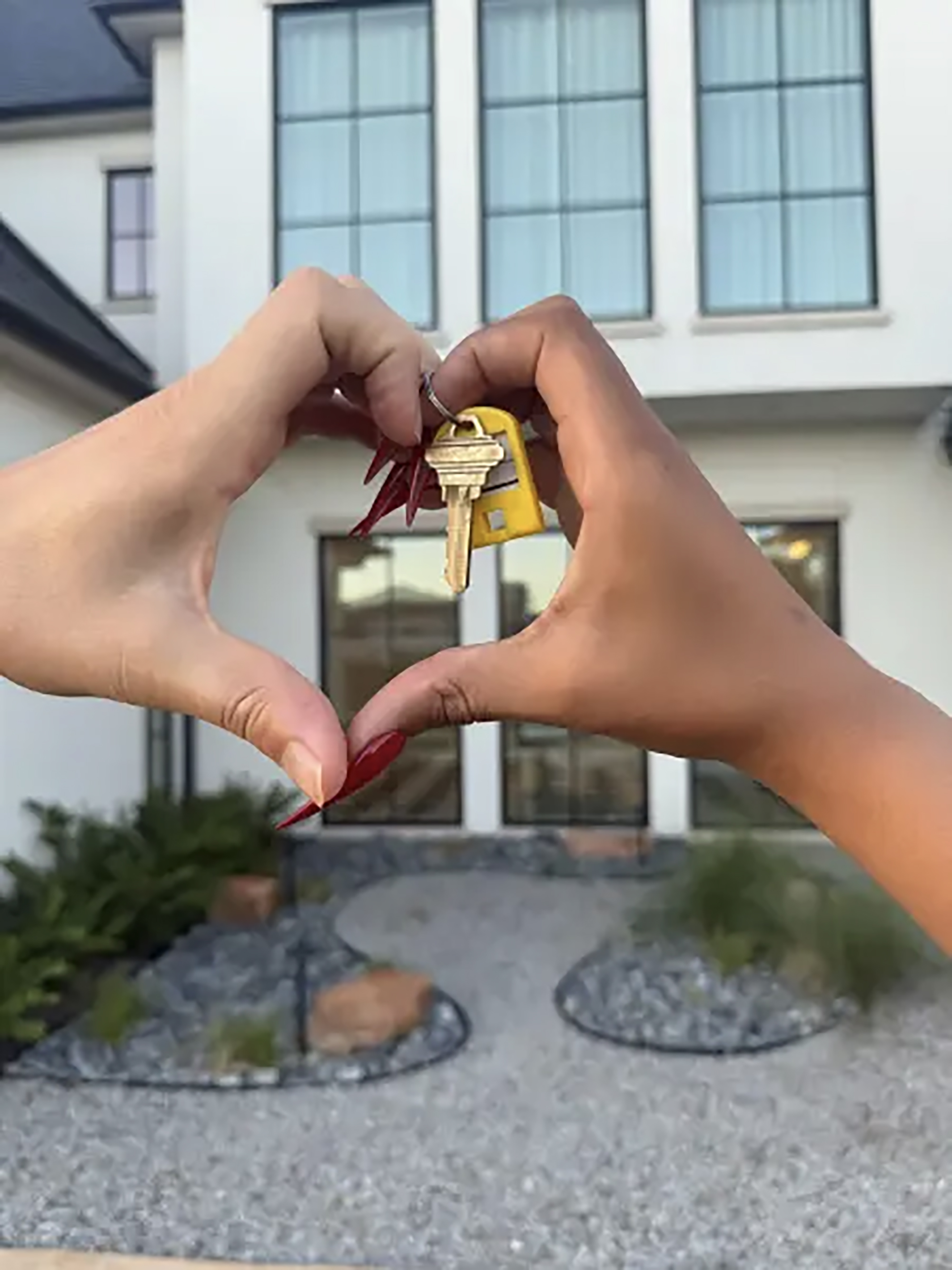 Two hands form a heart shape with house keys held in the middle, in front of a white house.