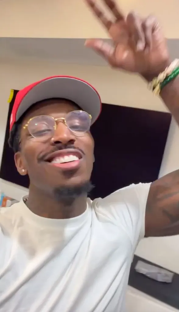 Pardison Fontaine smiling, wearing a red hat, gold glasses, a white t-shirt, and holding up two fingers.