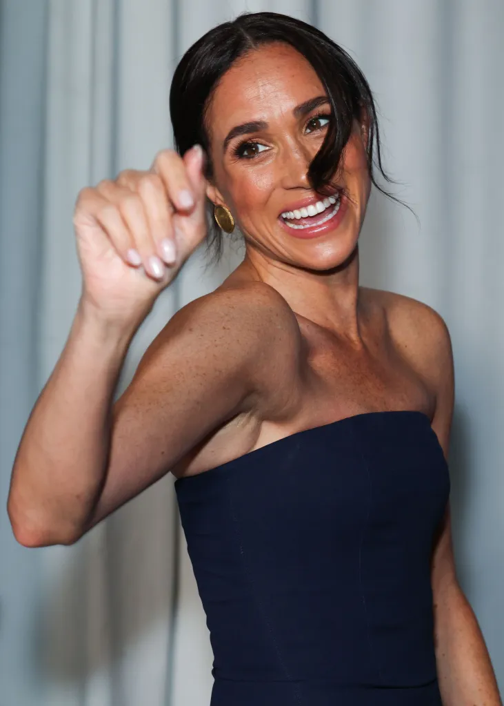 Meghan, Duchess of Sussex, smiling and gesturing with her right hand, wearing a dark blue strapless gown and gold earrings.
