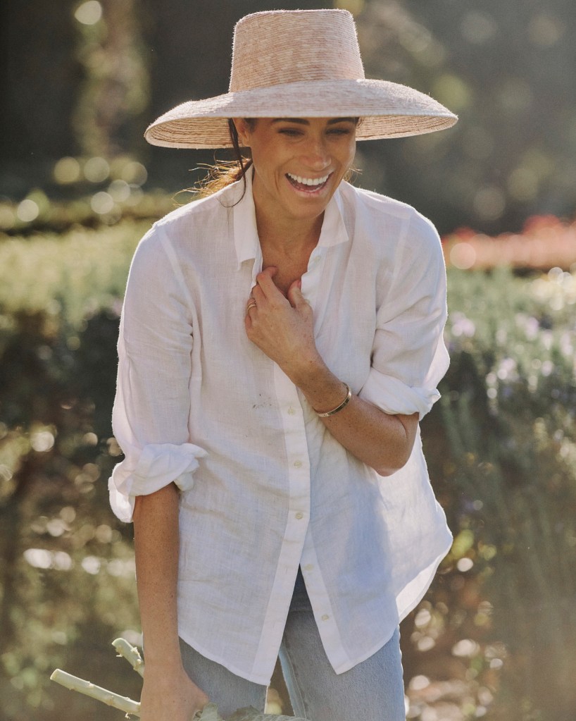 Meghan Markle laughing in her garden while wearing a straw hat and white shirt.