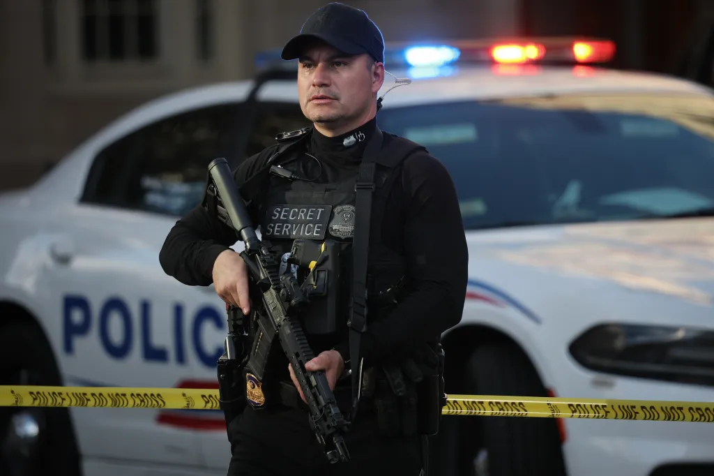 A U.S. Secret Service officer in tactical gear holds a rifle in front of a police car with flashing lights and crime scene tape.