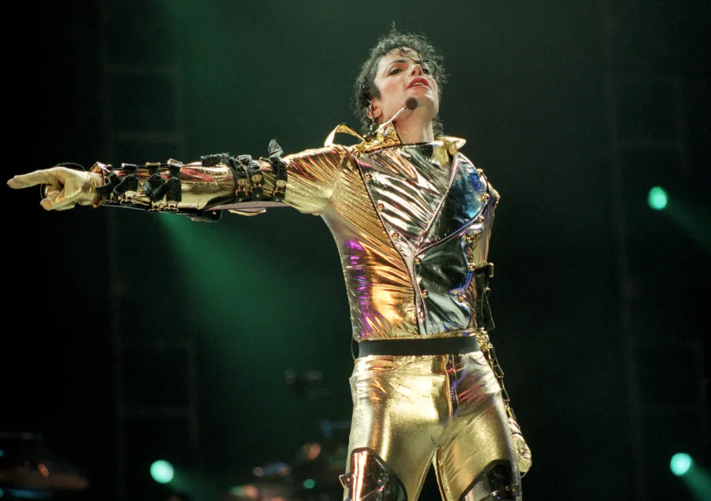 Michael Jackson performing in a gold and silver metallic costume with a microphone headset, pointing his gloved hand.