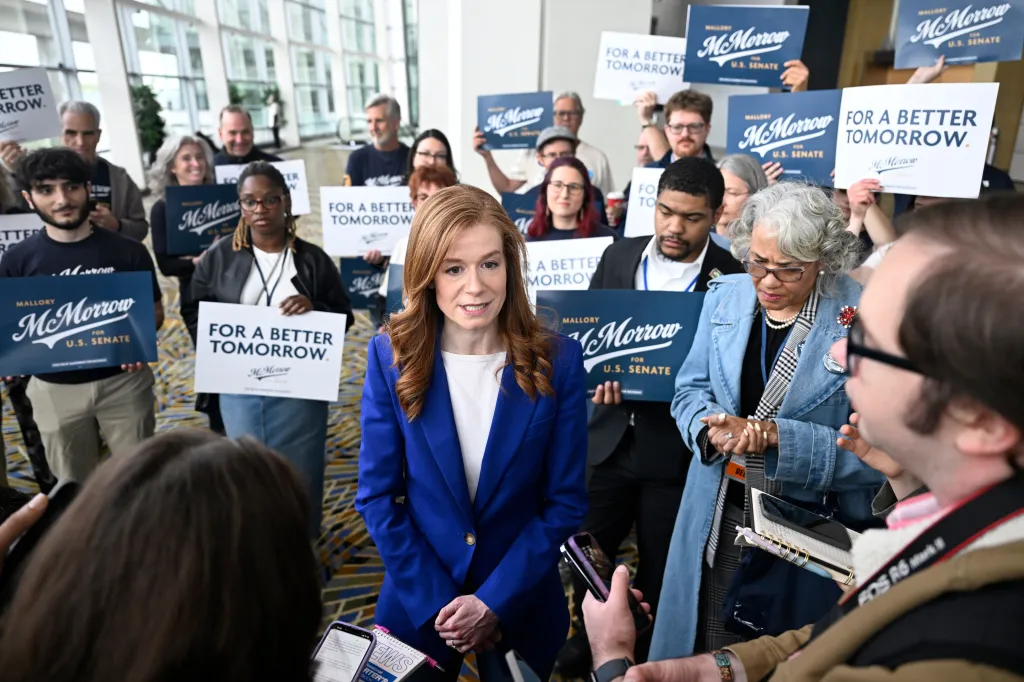 Michigan State Sen. Mallory McMorrow speaks to the media while surrounded by supporters holding campaign signs.