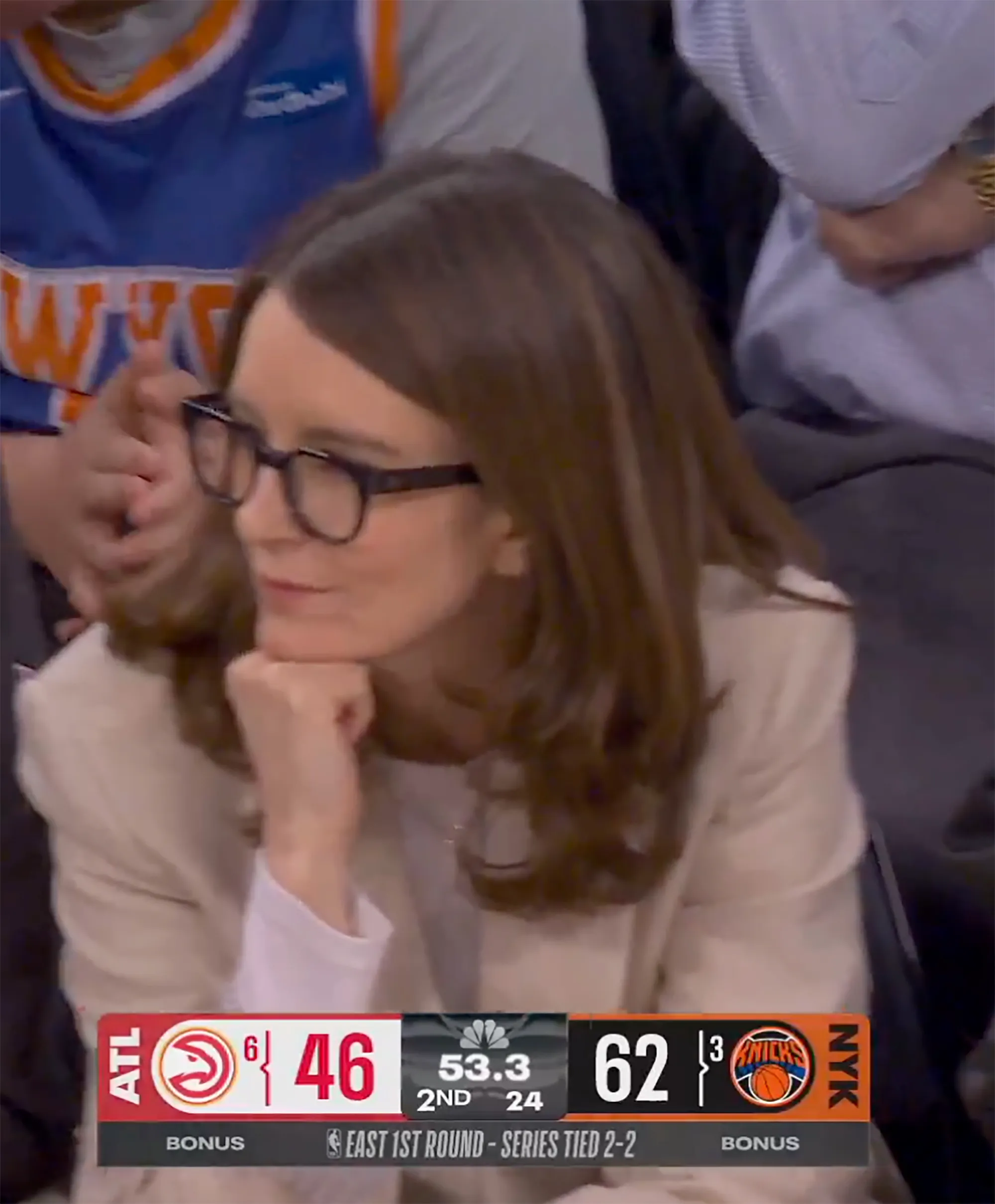 Tina Fey at a basketball game, wearing glasses and a light-colored jacket, with a game score overlay at the bottom.