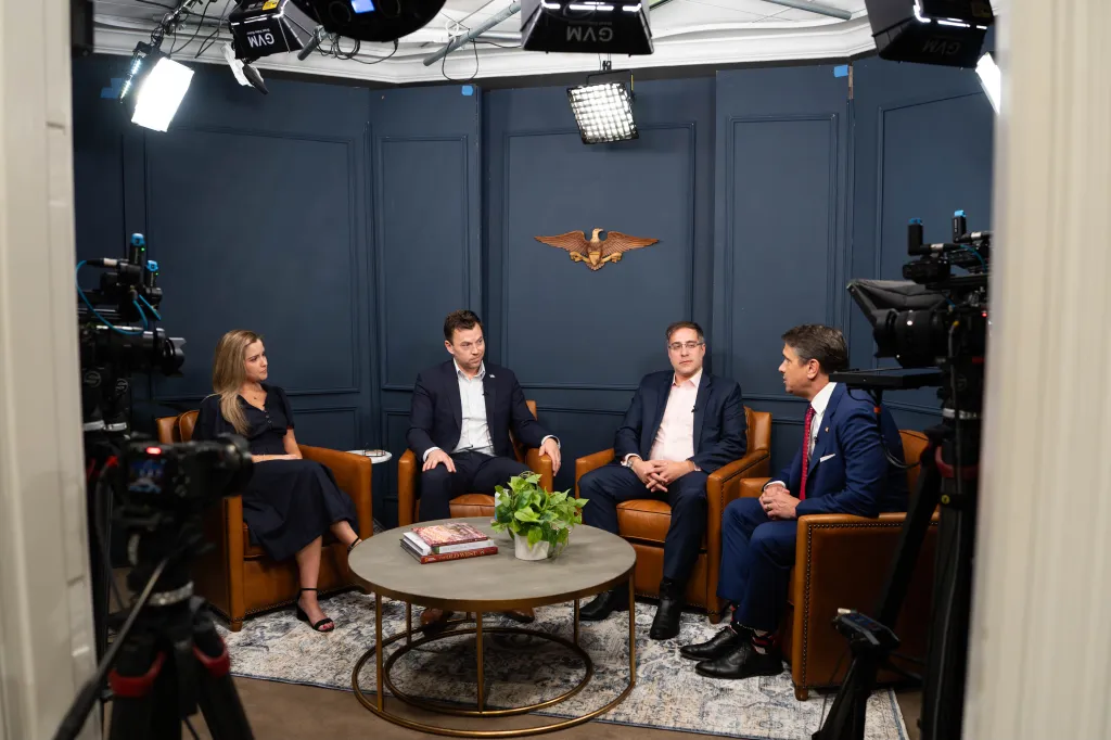 Todd Blanche (second from right) sitting in a brown leather chair, talking to other people in a broadcast studio set up.