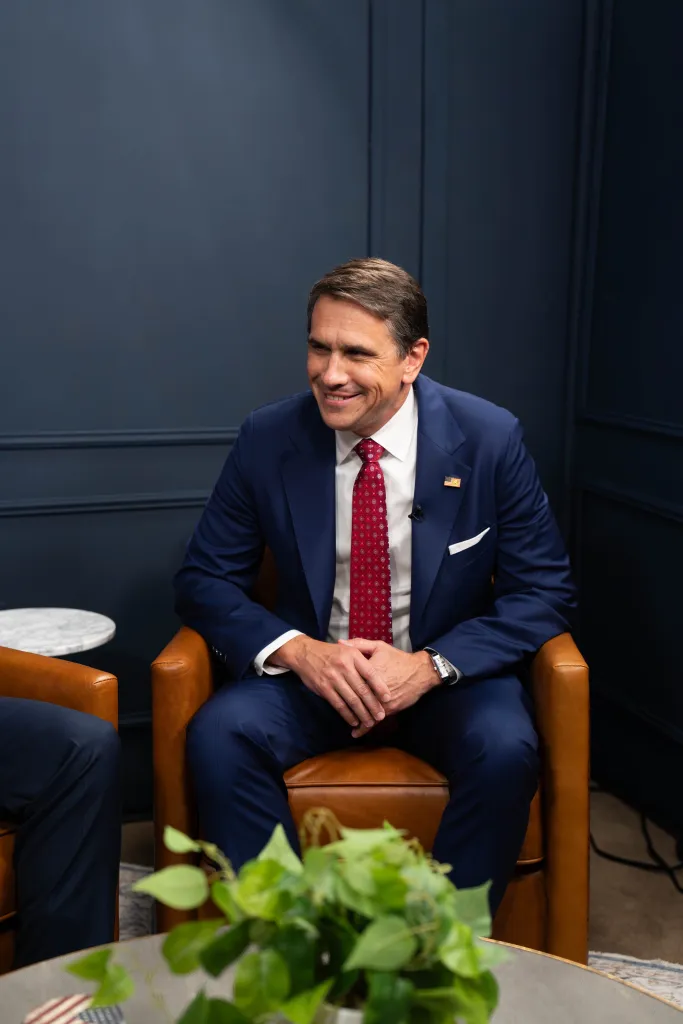 Todd Blanche, wearing a blue suit and red patterned tie, smiling while seated in a brown leather chair.