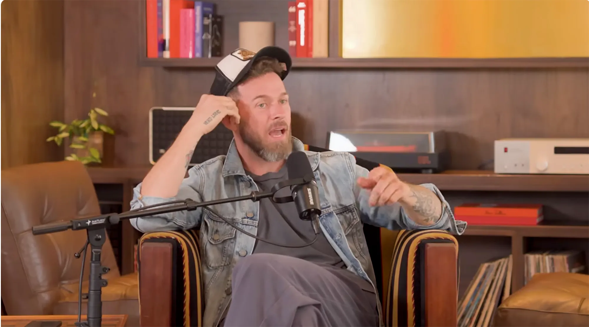 Artem Chigvintsev with a beard, wearing a denim jacket and a baseball cap, sitting in an armchair with a microphone in front of him.