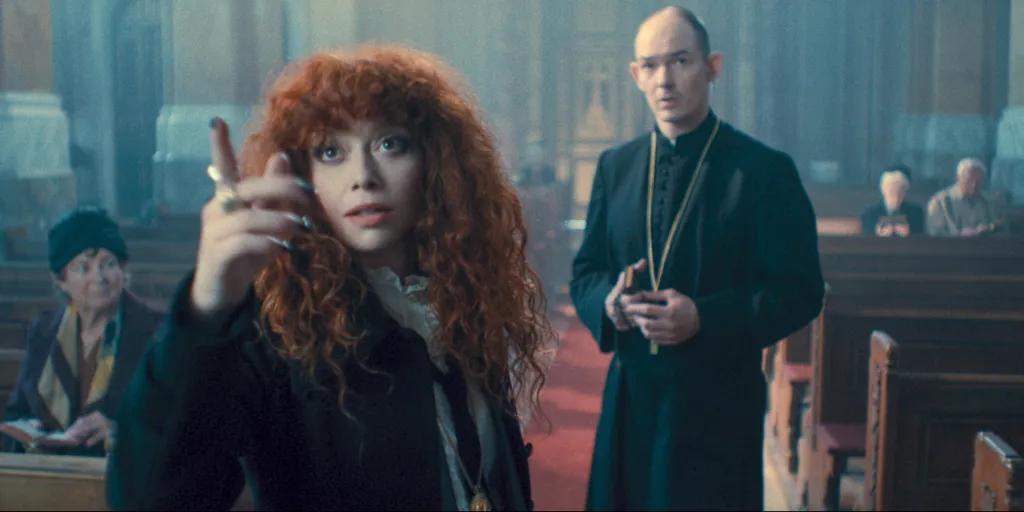 Natasha Lyonne , in a black jacket, points with her right hand while Akos Orosz in a black priestly cassock stands behind her in a church.