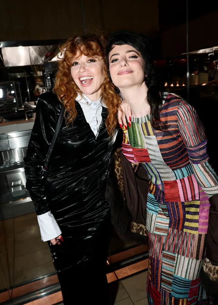Natasha Lyonne, in a black coat and white shirt, and Sarah Sherman, I a multi-colored striped dress, smiling and posing for a picture at an event.