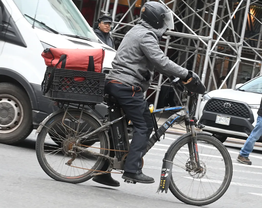 A food delivery worker on an e-bike, wearing a helmet, jacket, and dark pants, rides past a white van.