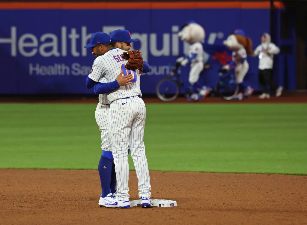 Two baseball players in white and blue striped uniforms hug on the field.