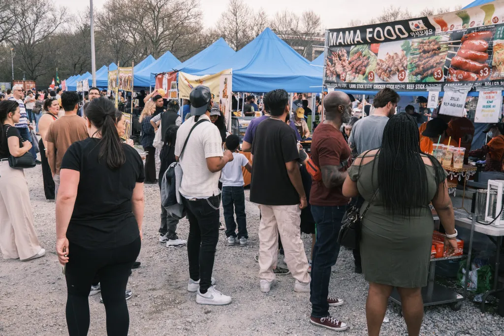 People gathered at the Queens Night Market, with a Mama Food stall offering various skewers and drinks.