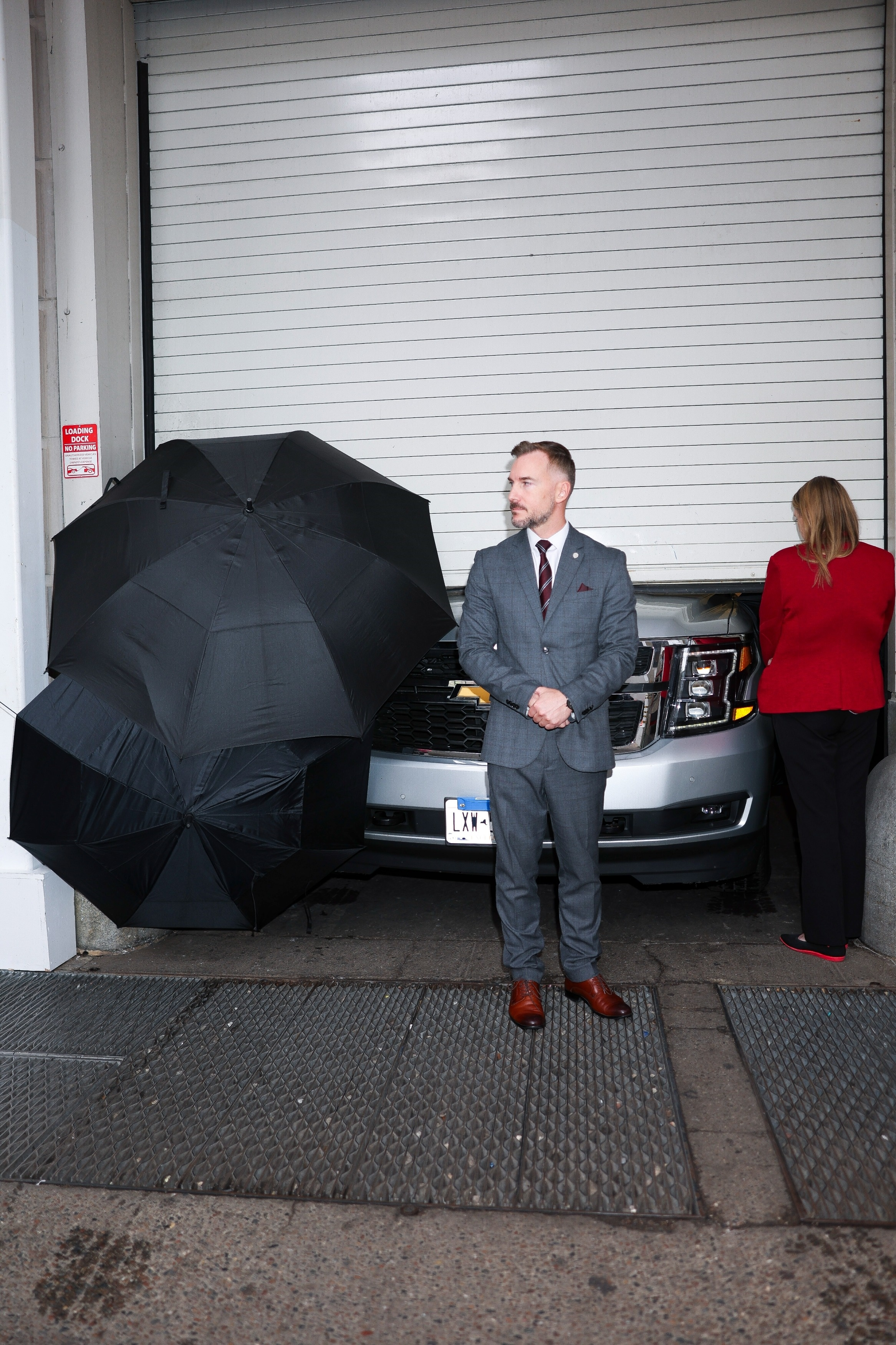 A man in a suit stands next to a car, an umbrella, and a woman in a red jacket, all in a garage.