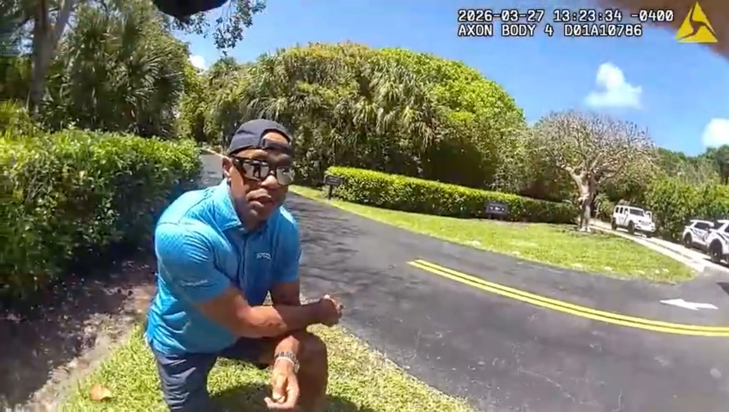 Tiger Woods sitting on his knee after a rollover accident, wearing sunglasses and a blue shirt, next to a road with a few white SUVs in the background.