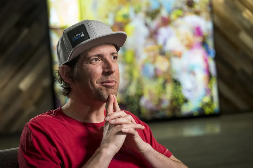 GoPro CEO Nick Woodman in a red shirt and gray hat, looking thoughtfully to the right with his hands clasped under his chin.