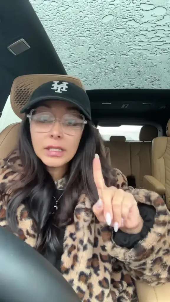 Nicole Polizzi in a leopard print coat, black hat, and glasses, pointing her finger.