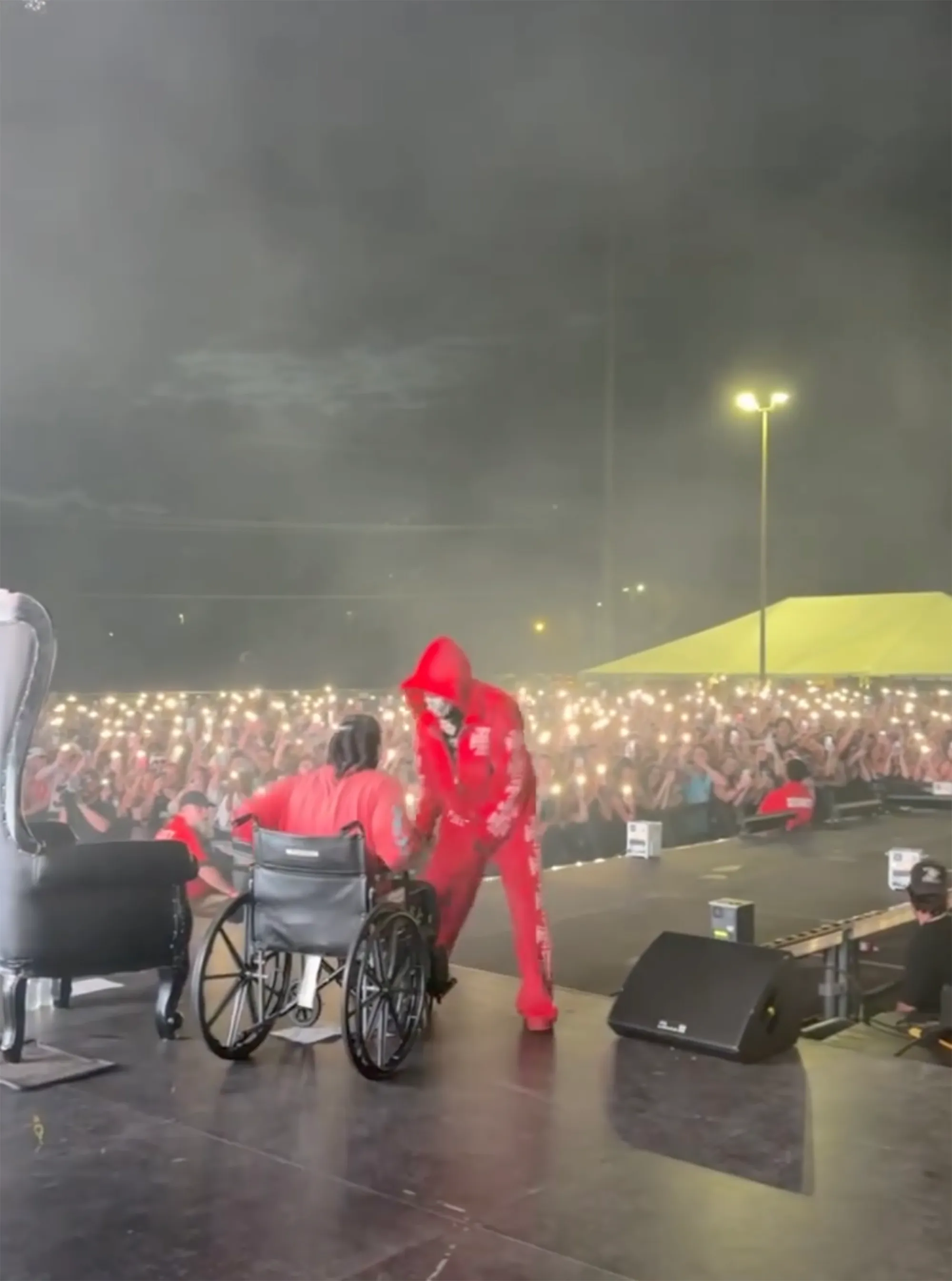 Offset exiting a wheelchair on stage, wearing a red jumpsuit, with a crowd of fans in the background holding up their phones.