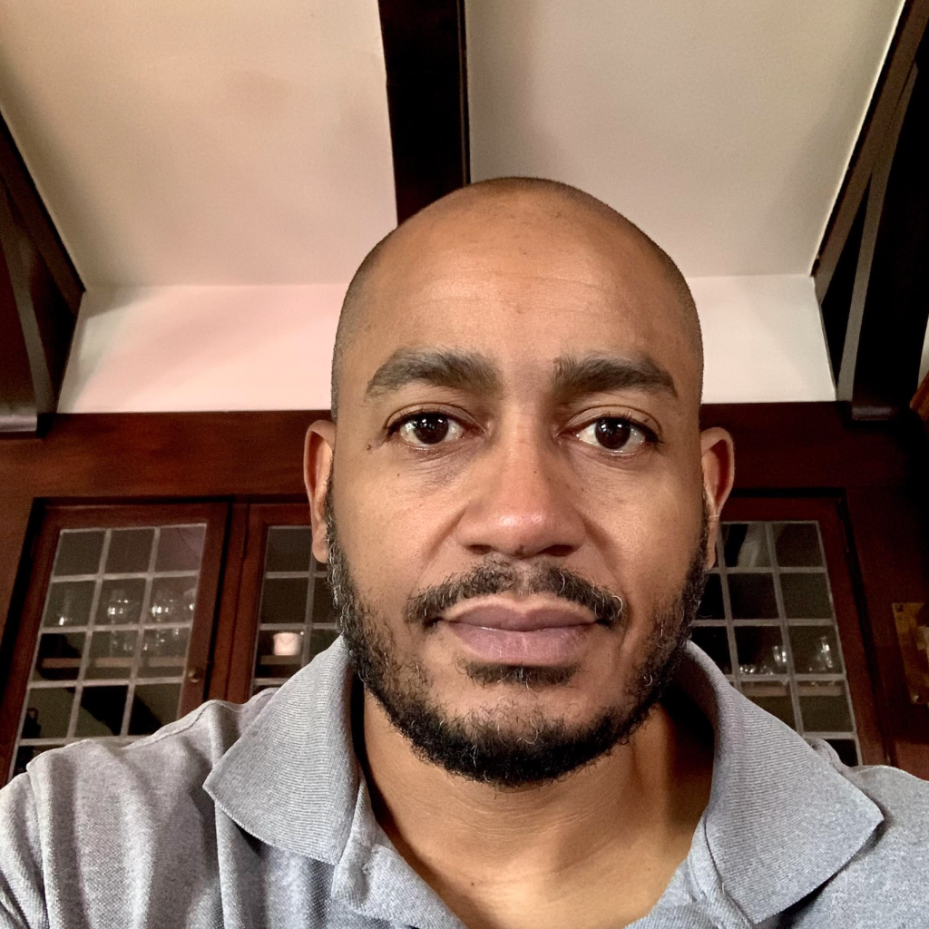 Headshot of Omar Farmer, a man with a shaved head and a dark beard, wearing a grey polo shirt.
